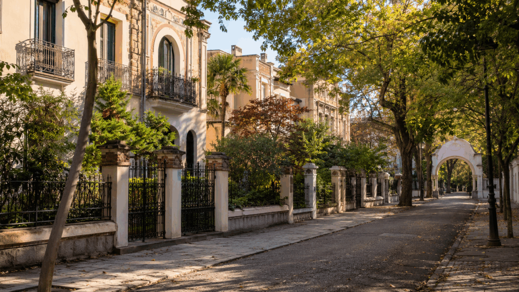 Calle residencial tranquila con casas históricas, jardines privados, verjas ornamentales, árboles y un arco de entrada al fondo, evocando el carácter singular de la Colonia de la Prensa en Carabanchel (IA)