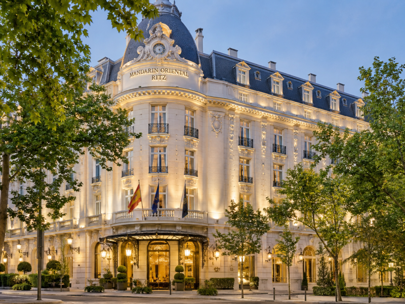 Fachada iluminada del hotel Mandarin Oriental Ritz Madrid al atardecer, con arquitectura clásica, cubierta de pizarra, balcones ornamentales, árboles en primer plano y entrada principal ajardinada. (IA)