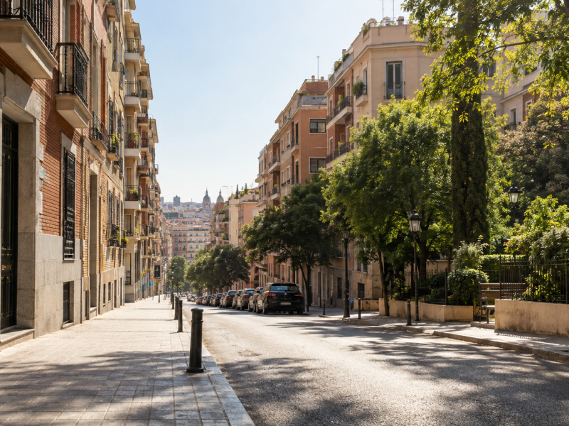 Calle urbana soleada con edificios residenciales, fachadas de ladrillo y piedra, árboles que proyectan sombra sobre la acera y vegetación abundante. (IA)