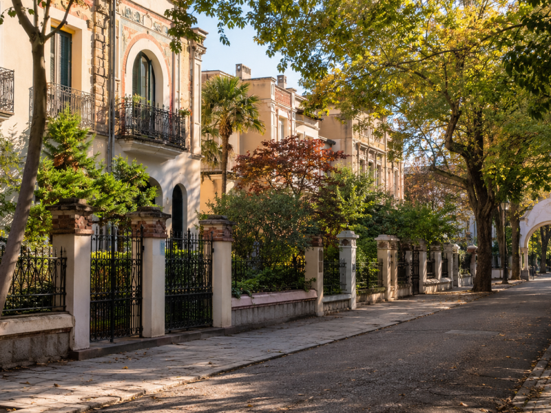 Calle residencial tranquila con casas históricas, jardines privados, verjas ornamentales, árboles y un arco de entrada al fondo, evocando el carácter singular de la Colonia de la Prensa en Carabanchel (IA)